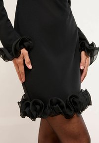 Černé šaty s dlouhými rukávy, které mají volánkové černé průhledné detaily na rukávech a spodním lemu. Hladká tkanina s vypasovaným střihem.