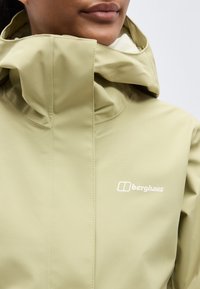 Άτομο που φοράει ανοιχτό πράσινο μπουφάν Berghaus με ψηλό γιακά και κουκούλα, με ορατό λογότυπο στο στήθος, τα χείλη και το πηγούνι φαίνονται μερικώς.