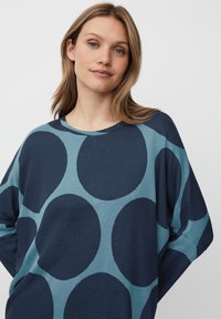 Tmavě modrý oversized svetr s světle modrým kruhovým vzorem. Měkká textura, široký výstřih a uvolněný střih s kleslými rameny.