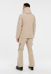 Beige waterdichte skijas met een capuchon, ritszakken en bijpassende beige skibroek met ritsen bij de enkels en zwarte skischoenen.