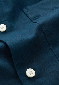 Gros plan sur un tissu bleu avec des boutons blancs et des coutures visibles sur un vêtement plié, probablement une chemise ou un chemisier.