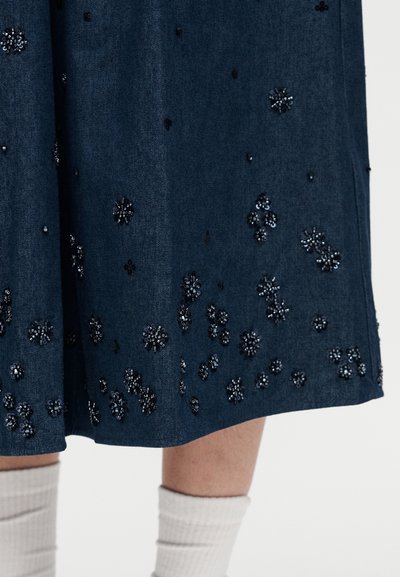 Σκούρο μπλε φούστα με σκορπισμένα μαύρα και ασημένια χάντρες σε σχέδιο λουλουδιών, φορεμένη με λευκές ριγωτές κάλτσες, εμφανιζόμενη από το μέσο του μηρού μέχρι το μέσο της γάμπας.