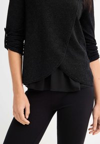 Top negro de manga larga con tejido texturizado, diseño en capas y dobladillo curvado. Se lleva sobre pantalones negros, con detalles de las mangas remangadas.