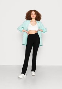 Blazer oversized verde menta, top cropped de malha branca, calças flared de malha pretas e ténis brancos. Fundo simples.