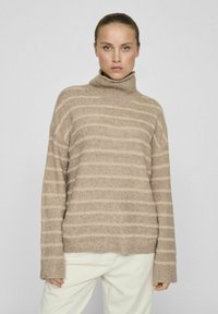 Bež pulover s turtleneck ovratnikom in horizontalnimi črtami v svetlejšem bež odtenku. Mehka tekstura, sproščen kroj in rebrasti manšeti. Usklajen z smetanastimi hlačami.