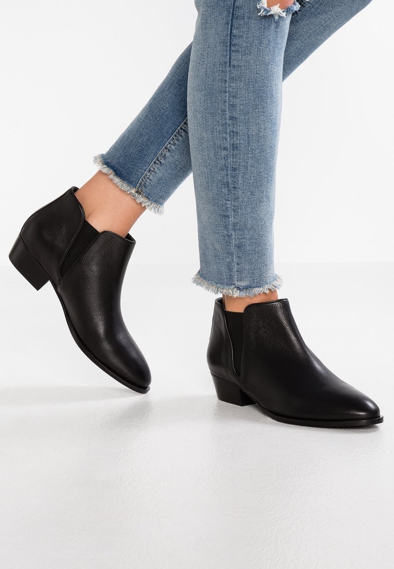 Bottines en cuir noir portées avec un jean bleu à ourlet effiloché sur fond blanc, un pied légèrement levé.