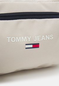 Sacchetto in tessuto beige con superficie testurizzata, con scritta bianca "TOMMY JEANS" e un piccolo logo con strisce rosse, bianche e blu. Chiusura con cerniera.