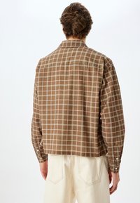 Camisa de cuadros marrón con cuello, mangas largas y puños abotonados; hecha de una tela suave con un diseño corto, combinada con pantalones blancos.