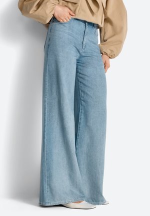 Persoon die lichtblauwe, wijde jeans met hoge taille draagt, beige top met lange mouwen en witte, puntige schoenen, poserend met één hand in de zak.