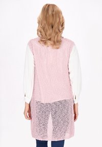 Lichtroze gebreide vest met een losse structuur en open rugontwerp, gedragen over een witte lange mouwtop en blauwe jeans.