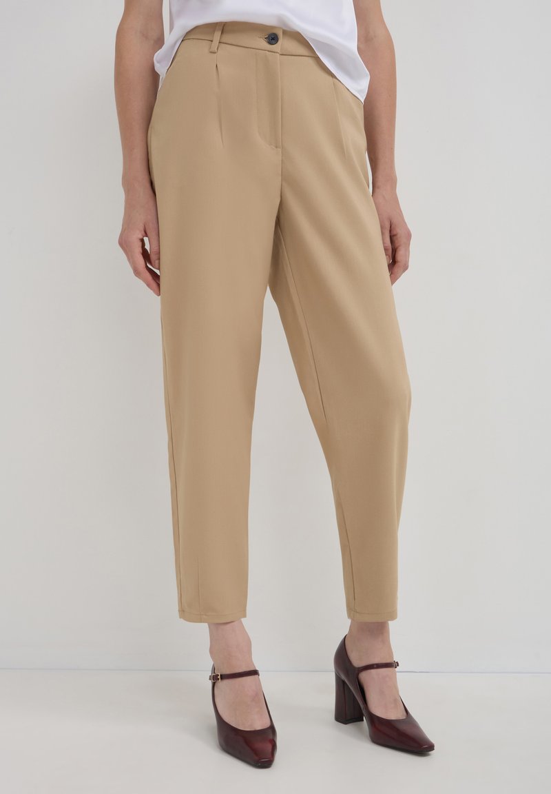 Pantaloni beige su misura con taglio dritto, caratterizzati da pieghe frontali e tasche laterali, abbinati a scarpe con tacco bordeaux.