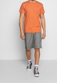 Camiseta de algodón naranja, estilo clásico con cuello redondo. Pantalones cortos grises enrollados en una tela texturizada. Zapatillas altas negras con cordones blancos.