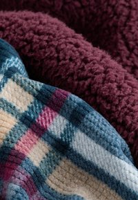 Kaksi päällekkäin olevaa peittoa: toinen syvän burgundin värinen fleece, toinen sinisessä ruutukuosissa, jossa on beige, vaaleanpunainen ja tummansininen korostus.