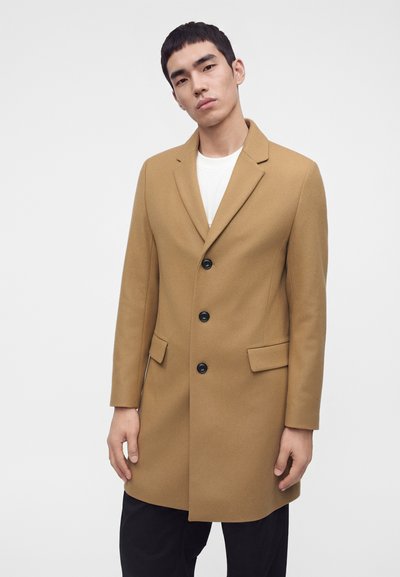 Homme portant un manteau en laine camel boutonné sur une chemise blanche et un pantalon noir, sur un fond clair uni.
