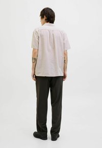 Chemise à manches courtes de couleur beige clair avec un col, détail de pli au dos, associée à un pantalon sombre et des chaussures, vue de derrière.
