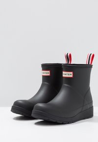 Bottes de pluie en caoutchouc noir avec une texture lisse, tige courte et poignées à rayures rouges et blanches. Étiquette de logo rouge sur les côtés.