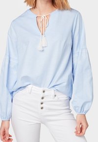 Helles blaues Baumwollbluse mit einem Schlüsselloch-Ausschnitt, gebunden mit Quasten, und Ballonärmeln mit dezenten Stickereien. Kombiniert mit weißen Jeans.