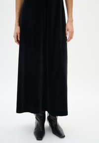 Frau in einem langen schwarzen Samtkleid und spitzen schwarzen Leder-Stiefeletten, stehend vor einem weißen Hintergrund.