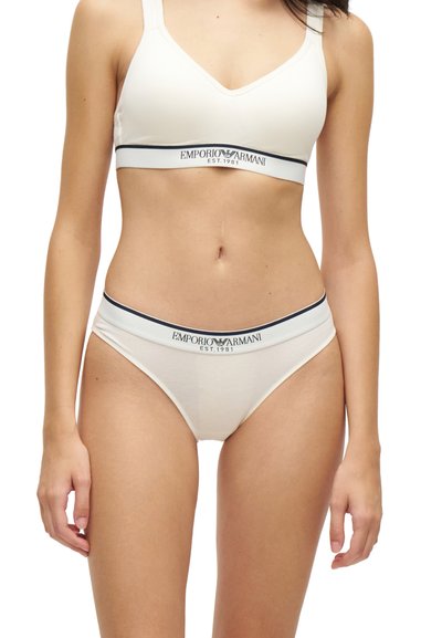 Femme portant un soutien-gorge blanc Emporio Armani et des sous-vêtements assortis avec un liseré noir et le logo de la marque sur les bandes, debout devant un fond blanc.