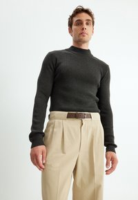 Dunkel gerippter Strickpullover mit hohem Kragen, kombiniert mit cremefarbenen, plissierten Hosen und einem braunen Ledergürtel mit einer silbernen Schnalle.