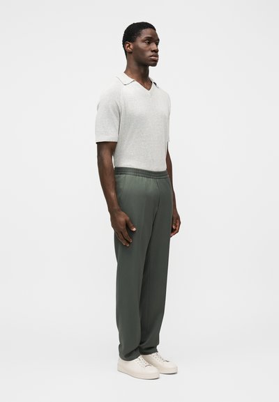 Ανοιχτό γκρι πλεκτό τοπ με κοντό μανίκι και V-λαιμόκοψη, συνδυασμένο με πράσινο παντελόνι με ελαστική μέση και λευκά αθλητικά παπούτσια, όλα σε απλό φόντο.