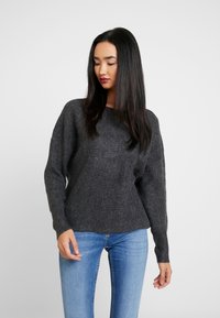 Pull en tricot gris foncé avec une coupe décontractée, des poignets côtelés et un large col bateau, associé à un jean bleu. Texture douce, design décontracté.