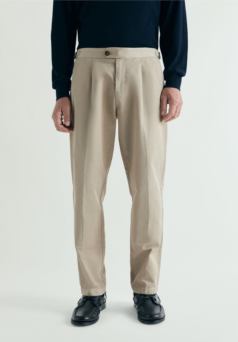 Hombre vestido con pantalones beige plisados con botón y ajustadores laterales, combinados con zapatos negros y una camiseta de manga larga azul marino sobre un fondo liso.