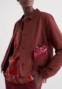 Pruunist puuvillast jakk, mis on varustatud nööbiga, vasakus taskus on mustriline punane taskurätt ja all on ruuduline särk.