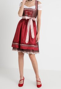 Robe dirndl traditionnelle avec une jupe en satin rouge ornée d'un ruban rose, un corsage brodé, une bordure en dentelle et un chemisier blanc à épaules dégagées.