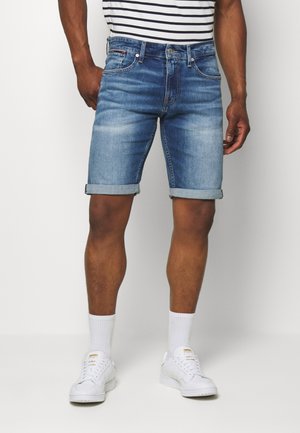 Homme portant un short en jean bleu roulé, des chaussettes blanches, des baskets blanches et une chemise rayée noir et blanc, debout sur un fond uni.