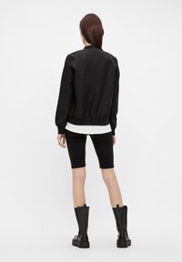 Mujer vistiendo una chaqueta bomber negra, pantalones cortos negros hasta la rodilla, camisa blanca y botas negras hasta la mitad de la pantorrilla, de pie de espaldas sobre un fondo blanco.
