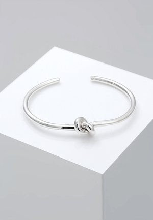 Bracciale a manetta aperto in argento con un dettaglio a nodo singolo al centro, esposto su una superficie bianca inclinata.