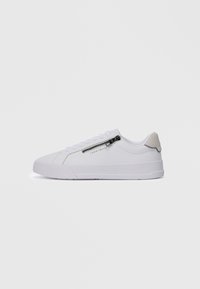 Hvit lav sneaker med siderom zipp og snøring, beige hæltab, og "Tommy Hilfiger" logo på siden.