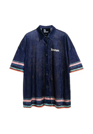 Hombre que lleva camisa azul marino y pantalones cortos con rayas rosas y verdes, diseño gráfico de ojo en la espalda, calcetines blancos y zapatos beige texturizados.