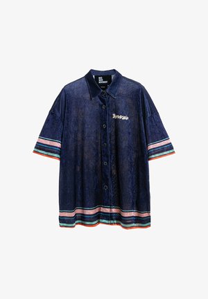 Hombre que lleva camisa azul marino y pantalones cortos con rayas rosas y verdes, diseño gráfico de ojo en la espalda, calcetines blancos y zapatos beige texturizados.