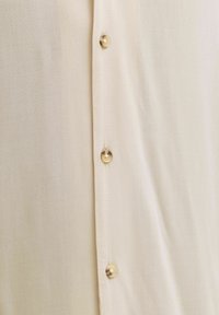 Gros plan sur un tissu de chemise beige avec trois boutons marron clair attachés le long de la couture centrale.