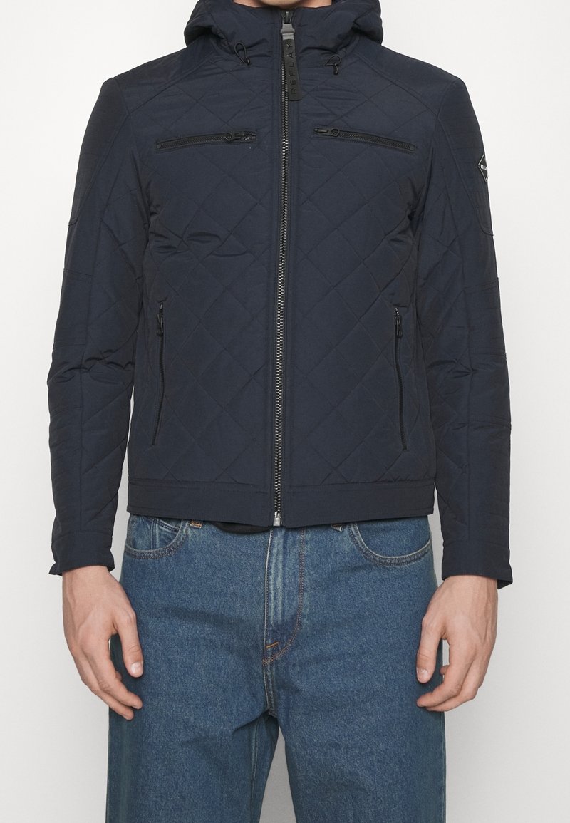 Veste matelassée bleu marine avec capuche, équipée d'une fermeture éclair frontale et de deux poches poitrine zippées. Texture lisse avec des détails de coutures subtiles.