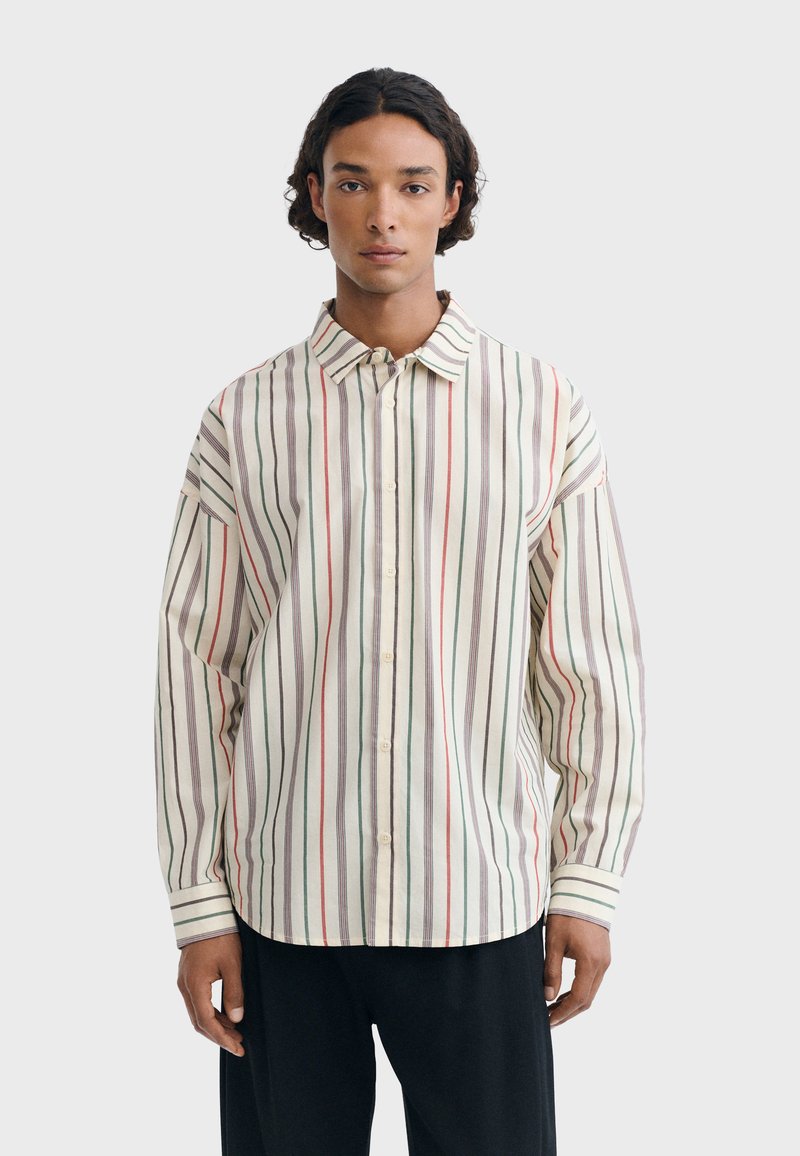 Jeune personne aux cheveux mi-longs et foncés portant une chemise ample à manches longues boutonnée avec des rayures verticales rouges, vertes et grises et un pantalon noir.