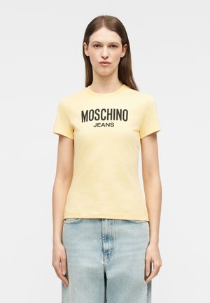 Mladá žena oblečená v svetložltom tričku Moschino Jeans a svetlomodrých džínsových nohaviciach na jednoduchom svetlom pozadí.