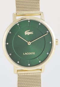 Montre avec bracelet en maille dorée et cadran vert mat, présentant un logo de crocodile et des accents étincelants à chaque repère horaire.