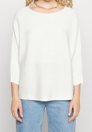 Maglione bianco a collo alto con lavorazione a maglia, scollatura ampia e maniche a tre quarti. Abbinato a jeans in denim azzurri chiari. Collana in catena dorata.