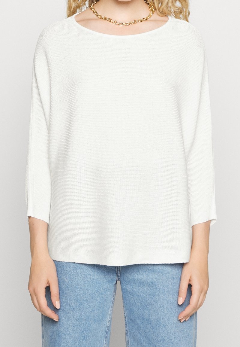Weißer Pullover mit strukturiertem Strick, weitem Ausschnitt und dreiviertellangen Ärmeln. Kombiniert mit hellblauen Jeans. Goldene Kettenhalskette.