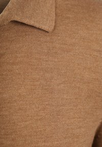 Bruine gebreide trui met een geribbelde textuur en een puntige kraag. De stof lijkt zacht en lichtgewicht, met een gladde, gelijkmatige afwerking.