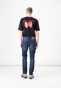 T-shirt noir avec un graphic coloré de pomme et le texte "dieselcore" dans le dos, associée à un jeans bleu et des baskets grises.