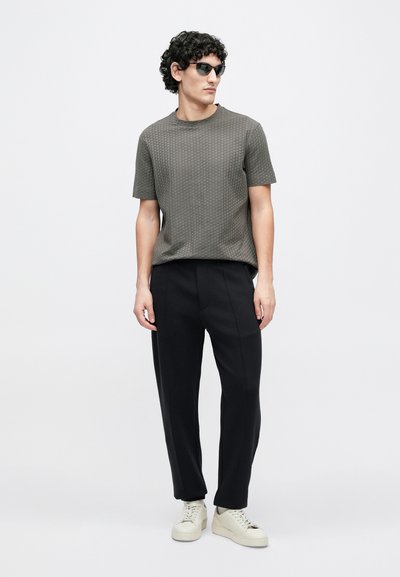 Pilkas tekstūruotas trumpomis rankovėmis marškinėliai, derinami su juodomis pritaikytomis kelnėmis ir baltais sportiniais bateliais, stovint prieš paprastą baltą foną.