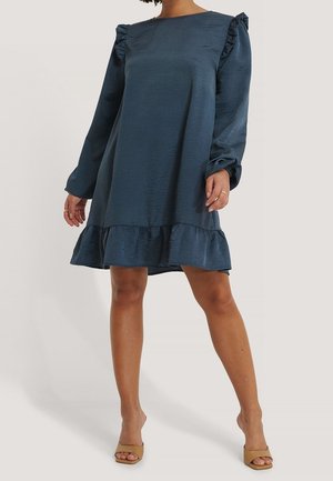 Kvinde iført en knælængde mørkeblå kjole med flæser på skuldrene og kanten, kombineret med beige åben-tå hæle, stående på en lys baggrund.