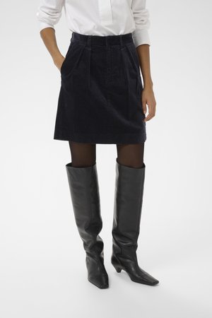 Jupe en velours côtelé bleu marine foncé avec des plis à la taille, associée à des bottes noires en cuir montant jusqu'aux genoux, dotées d'un bout pointu et de talons blocs.