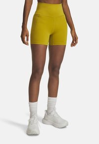 Gele high-waisted shorts met een gladde textuur, zijzakken en contrasterende stiksels, gecombineerd met witte sportschoenen en sokken.