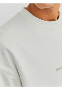 Ljusgrå sweatshirt med ribbad rundhalsringning. Har minimal sömnad och en liten präglad logotyp i en kontrasterande färg.
