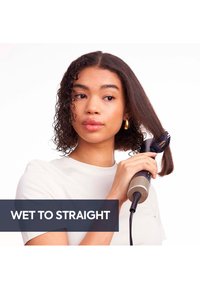 Junge Frau mit lockigem Haar benutzt ein Haarstylinggerät, um eine Strähne ihres Haares zu glätten, trägt ein weißes Hemd und goldene Creolen.
